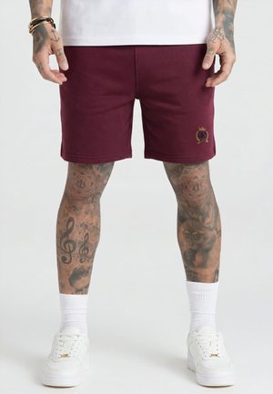 Hombre con pantalones cortos color burdeos con logo de escudo, calcetines blancos y zapatillas blancas, mostrando piernas y manos tatuadas contra un fondo liso.