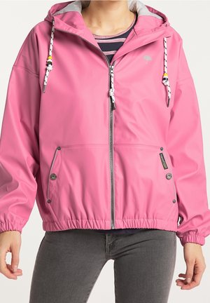Personne portant une veste de pluie rose à capuche zippée avec poches avant, poignets élastiques et cordons blancs sur une chemise rayée et un pantalon gris.