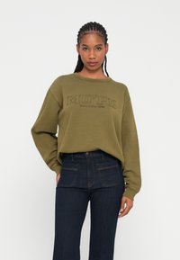 Felpa verde oliva con un logo testurizzato "MO’PD" e "Marc O'Polo DENIM" ricamato. Abbinata a jeans in denim blu scuro a vita alta.