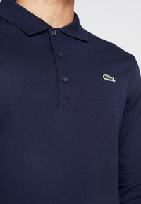 Námorně modrá polo košile s dlouhým rukávem, texturovaná tkanina, tříknoflíková léga a vyšívané zelené logo krokodýla na levé hrudi.