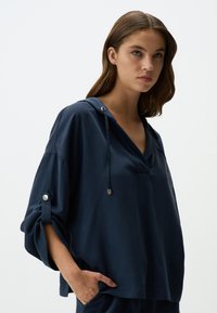 Pullover blu navy con cappuccio, maniche ampie e dettagli in metallo argentato. Tessuto morbido e vestibilità rilassata, con scollatura a V e tasche laterali.