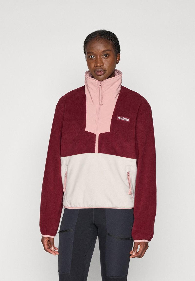 Kvinne iført en fargeblokk fleece-pullover i burgunder, rosa og krem, som står mot en enkel lys bakgrunn.