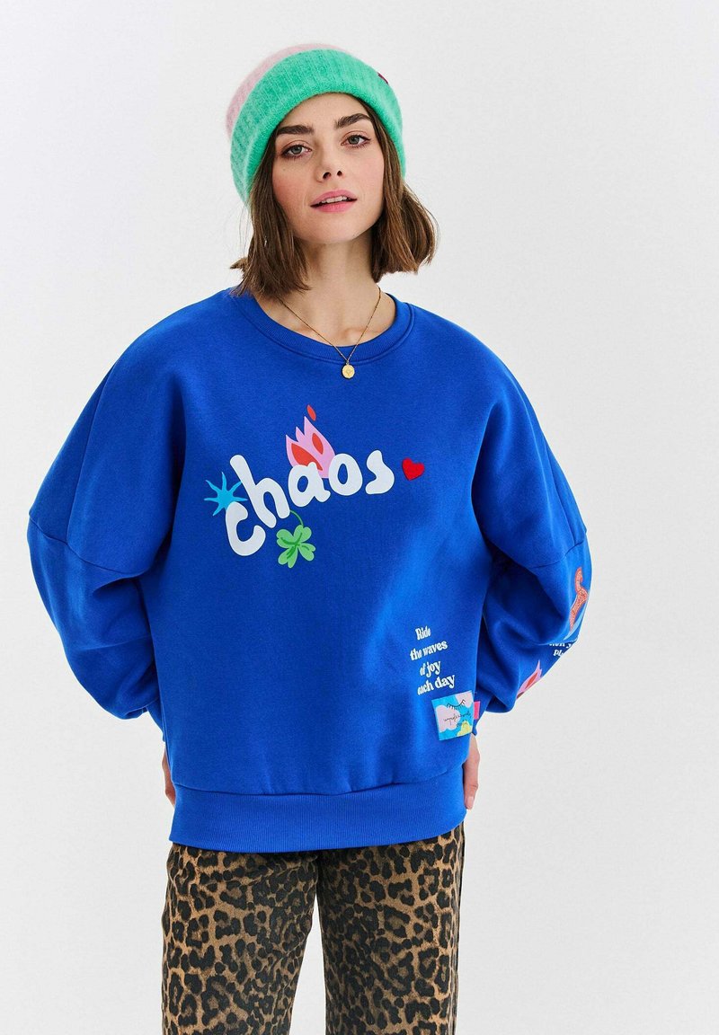 Modrá mikina s farebnými grafikami vrátane slova "chaos", nosená s nohavicami s leopardím vzorom. Mäkkučká tkanina a voľný strih.