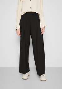 Černé široké kalhoty s sklady a páskem v pase. Spárovány s off-white blůzou s knoflíkovými manžetami a uvolněným střihem.