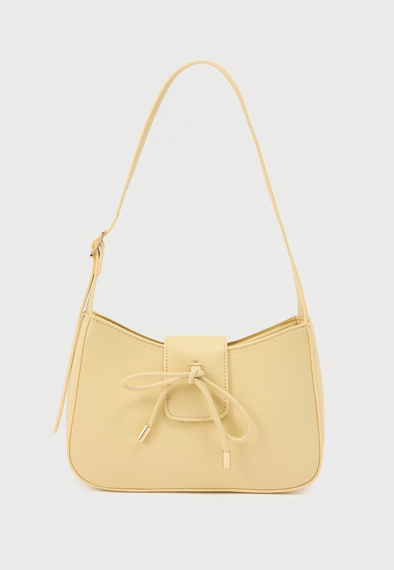 Μικρή μπεζ τσάντα ώμου με ρυθμιζόμενο λουρί και μπροστινό καπάκι δεμένο με λεπτό κορδόνι που τελειώνει σε μεταλλικά μύτα, σε λευκό φόντο.