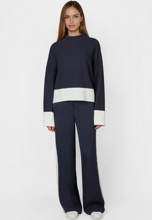 Young woman wearing a navy blue sweater with white cuffs and hem, paired with matching wide-leg pants and white sneakers, standing against white background.