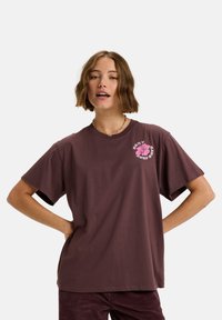 Bruine katoenen t-shirt met korte mouwen, met een roze bloemenafbeelding op de linkerborst. Rechte snit en zachte textuur.