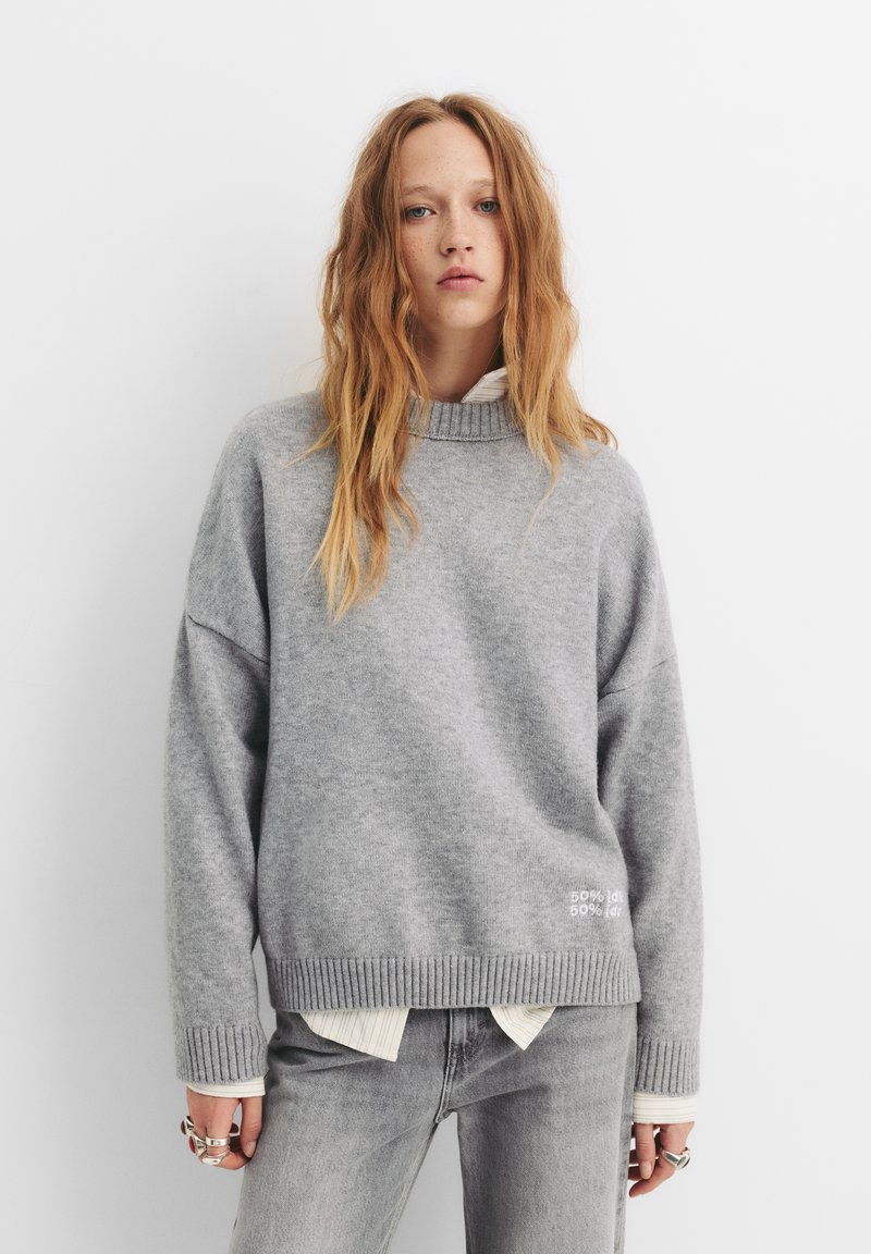 Maglione grigio oversize con collo e orlo a coste, realizzato in un materiale morbido. Presenta un dettaglio di testo bianco sulla parte anteriore inferiore. Abbinato a jeans chiari.