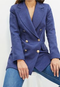 Blazer croisé à fines rayures bleues avec accents de boutons dorés, doté d'un col cranté et d'épaules structurées, associé à un jean en denim.