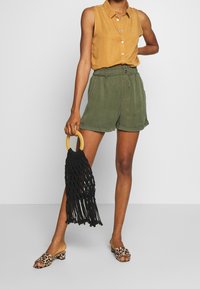 Khaki-shorts och en senapsgul ärmlös tröja, kombinerat med en svart fransad handväska och leopardmönstrade klackar med öppen tå.