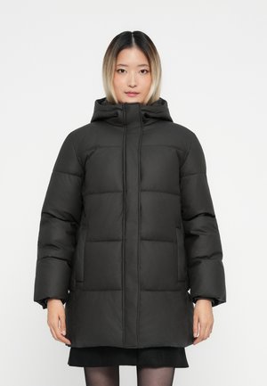 Femme portant un manteau d'hiver noir rembourré avec capuche, se tenant devant un fond blanc uni.