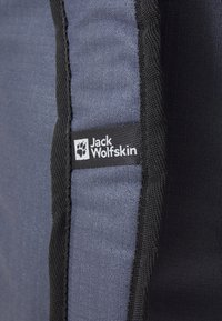 Etichetta Jack Wolfskin su tessuto grigio e nero, che mostra una texture e una cucitura resistenti. Presenta il logo con il dettaglio di una impronta di zampa.