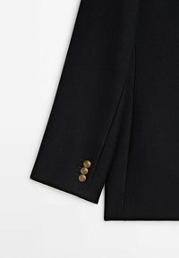 Manche de blazer noire sur mesure avec trois boutons décoratifs dorés sur le poignet, sur fond blanc.