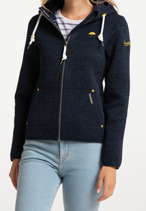 Femme portant un sweat à capuche zippé bleu foncé avec des cordons blancs et un logo parapluie jaune, associé à un jean bleu clair et une chemise rayée.