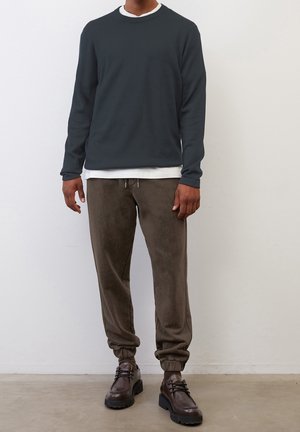 Dunkelgraues Langarmshirt, das über einem weißen Rundhalsshirt getragen wird, kombiniert mit braunen Velour-Jogginghosen und braunen Schnürstiefeln mit dicken Sohlen.