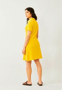 Keltainen polo-tyylinen mekko lyhyillä hihoilla ja levenevällä helmalla. Malli on pukeutunut ruskeisiin sandaaleihin, ja hänet on kuvattu takaapäin neutraalilla taustalla.