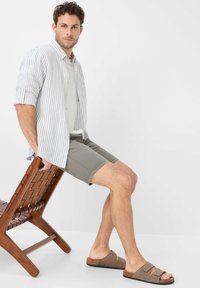 Camicia a righe leggera e con bottoni; pantaloni corti grigi; sandali marroni a scivolo con doppie cinghie; seduto su una sedia di legno con design intrecciato.
