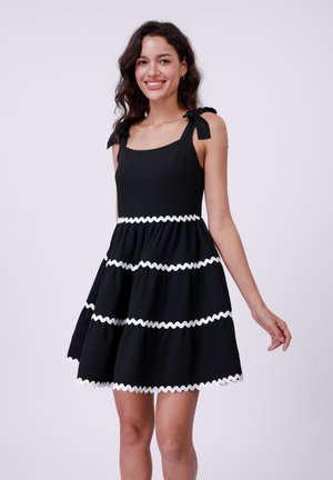 Robe noire sans manches avec un décolleté carré, ornée de garnitures blanches ondulées sur trois niveaux, et de liens sur les bretelles.