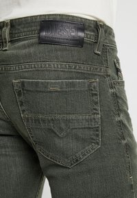 Gröna jeans i denim med strukturerad yta, utrustade med en svart läderlogotyp, dubbla bakfickor och gula sömnadsdetaljer.