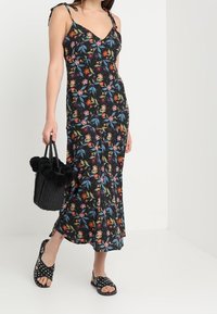 Svart blommig maxiklänning med justerbara axelband, prydd med färgglada blommor och gröna löv. Matchas med en strukturerad svart väska och sandaler med prickar.