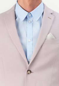 Απαλή ροζ σακάκι με πέτο, κλείσιμο με έναν κουμπί και λευκή τσέπη; φορεμένο πάνω από ένα ανοιχτό μπλε πουκάμισο με κουμπιά.