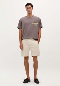 Grå kortærmet t-shirt med gult "SCALPERS" logo; parret med cremefarvede shorts med lommer og knaplukning. Sorte sko.