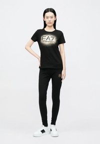 Sort t-shirt og matchende bukser med "EA"-logo i guldtryk. Outfitet er lavet af blødt stof og suppleres med hvide sneakers.