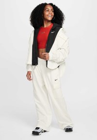 Biała oversize'owa kurtka z czarnymi akcentami, czerwona krótka bluzka, luźne białe spodnie i srebrno-czarne sneakersy. Zawiera logo Nike i luźny krój.