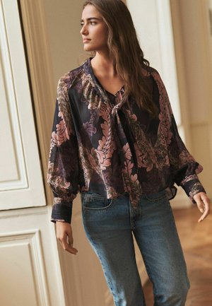 Blus med blommönster i mörka färger med puffärmar, knuten med en snörning vid halsen, i kombination med ljusblå jeans. Slät textur.