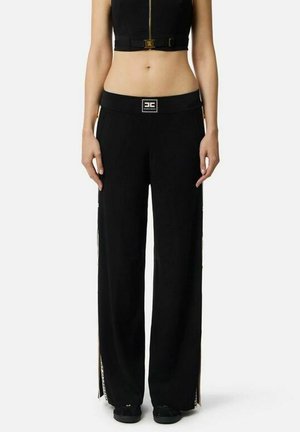 Donna che indossa pantaloni neri a gamba larga con una fascia elastica con logo e un top corto nero, in piedi davanti a uno sfondo semplice.