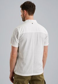 Camisa de color claro de manga corta hecha de tejido texturizado, con cuello, costura en la parte posterior y un pequeño logotipo cerca del cuello.