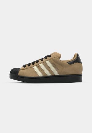 Tan suede sneaker with black rubber toe cap, three white side stripes, black laces, and a black leather heel accent.