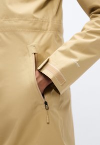 Μπεζ αδιάβροχο μπουφάν με μπροστινή τσέπη με φερμουάρ, ρυθμιζόμενο μανσέτα με Velcro και σήμανση "DRY VENT" στο μανίκι. Ομαλή υφή.