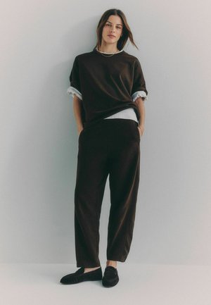 Nő áll egy sima fal előtt, laza sötétbarna ruhában, felgöngyölített ujjakkal, fekete mokaszinokban, kezével a zsebében.