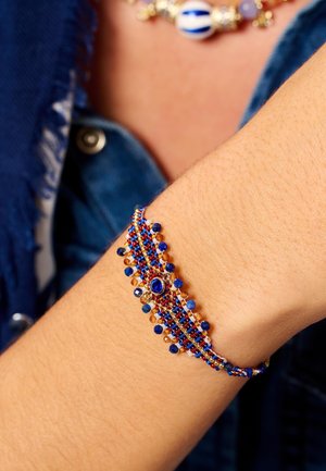 Bracelet en perles présentant des teintes bleues, rouges et dorées avec des motifs de perles complexes et un accent central en pierre bleue, posé sur un poignet.