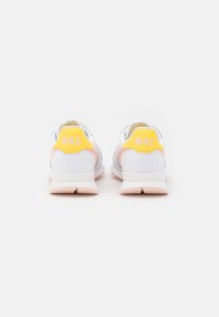 Tenisky Nike s bílým koženým svrškem a růžovými detaily, s žlutou patní štítkem s nápisem „NIKE“ v bílém textu. Růžová gumová podrážka.