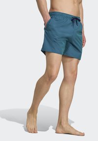 Bañadores de hombre en verde azulado con rayas verticales blancas, cinturilla elástica, cordón y bolsillos laterales. Tejido suave con un corte holgado.