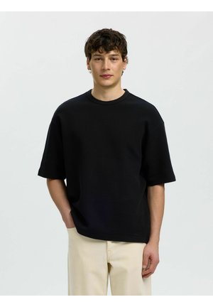 Fiatal férfi göndör hajjal, fekete rövid ujjú ingben és bézs nadrágban, áll egy egyszerű világos háttér előtt.