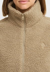 Veste en polaire beige avec un col montant, une fermeture éclair intégrale et une surface texturée. Présente un logo embossé subtil sur la poitrine.
