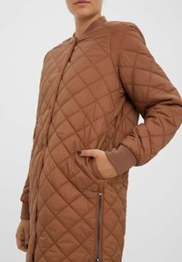 Veste longue marron matelassée avec une poche zippée, des poignets côtelés et un col arrondi, fabriquée dans un matériau lisse et léger.