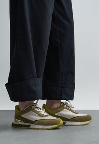 Groen- en beige sneakers met suède en mesh materialen, voorzien van een gestructureerd ontwerp en een metalen detail op de hiel. Draagt bij een donkere broek.