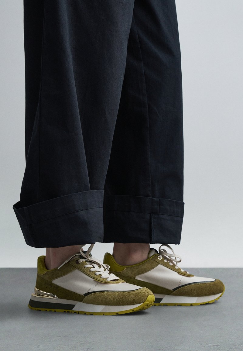 Groen- en beige sneakers met suède en mesh materialen, voorzien van een gestructureerd ontwerp en een metalen detail op de hiel. Draagt bij een donkere broek.