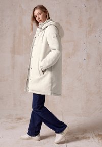 Witte fleece-voering parka met een capuchon, drukknoopsluitingen en zijzakken, gecombineerd met donkerblauwe spijkerbroek en witte sneakers. Textuur en warme uitstraling.