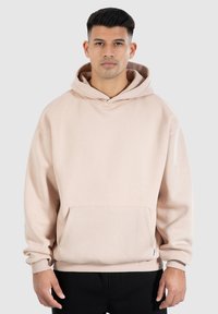 Hoodie rose clair avec un cordon de serrage, poche frontale, poignets et ourlet côtelés. Tissu doux avec une coupe décontractée, modèle portant un pantalon noir.