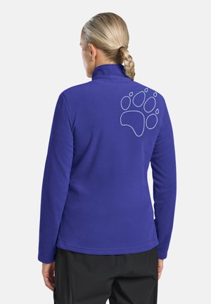 Persoon met gevlochten haar die een blauw fleecejack draagt met een grote omrande pootafdruk op de bovenrug, staand tegen een witte achtergrond.