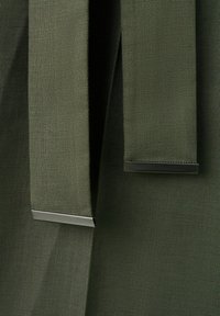 Nærbillede af olivengrønt stof med to hængende stropper, hver afsluttet med en skinnende metalspids.