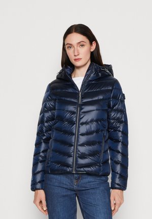 Veste rembourrée bleue marine avec capuche, affichant une finition brillante, un motif matelassé, une fermeture éclair à l'avant et des poches latérales. Portée par-dessus un haut blanc.