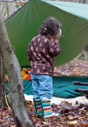 Child wearing a brown sweatshirt with owl patterns, light blue pants, and colorful striped rain boots, standing under a green tent.