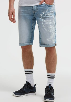 Mann trägt hellblaue aufgerollte Jeansshorts mit weißem Tiger-Print und Schriftzug, weiße Socken mit schwarzen Streifen und schwarze Sneaker.