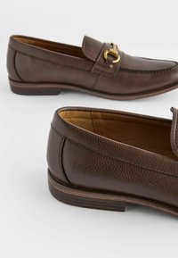 Mocassins en cuir marron avec une finition texturée, une boucle en ton doré en accent et un talon légèrement surélevé. Doublure intérieure lisse visible.